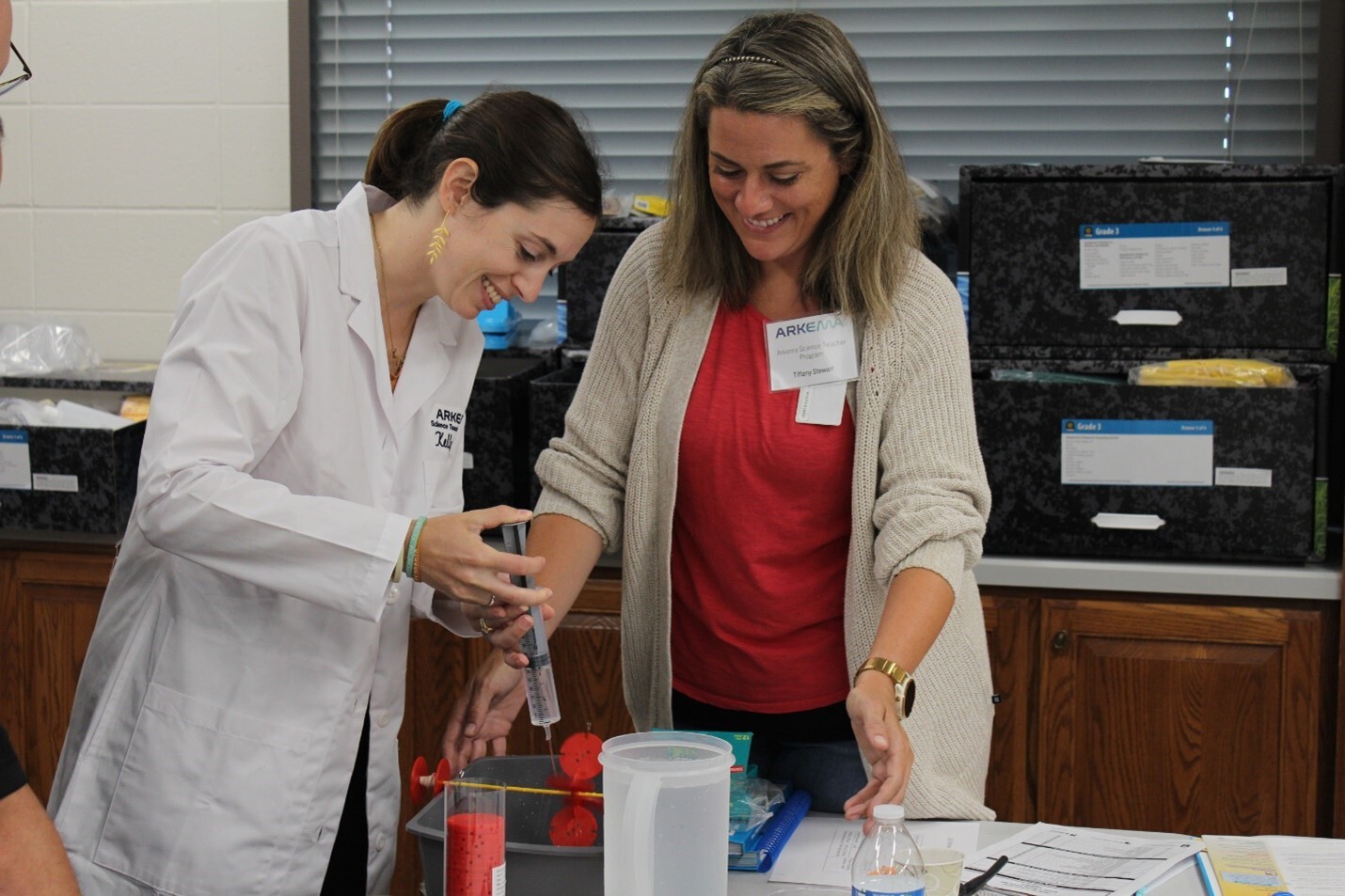 Two women smiling, one in a lab coat, working together on a science experiment.